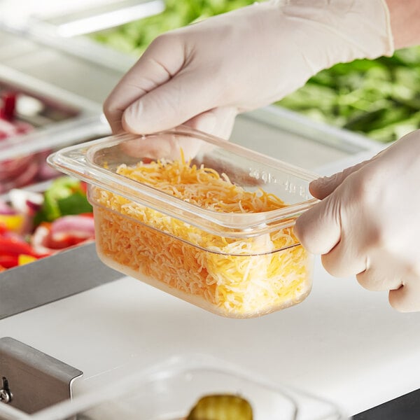 A person in gloves holding a clear polycarbonate food pan of shredded cheese.