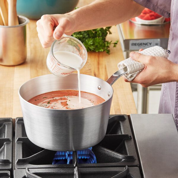 A person pouring milk into a Vollrath Wear-Ever sauce pan on a stove.