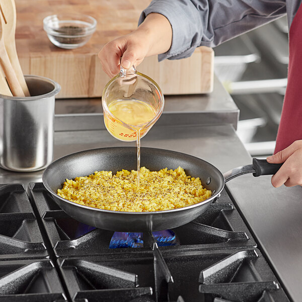 A person pouring liquid from a measuring cup into a Vollrath stainless steel non-stick fry pan on a stove.