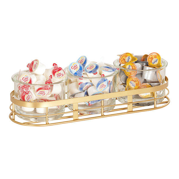 A Cal-Mil gold metal coffee condiment organizer with three glass jars filled with candy on a counter.