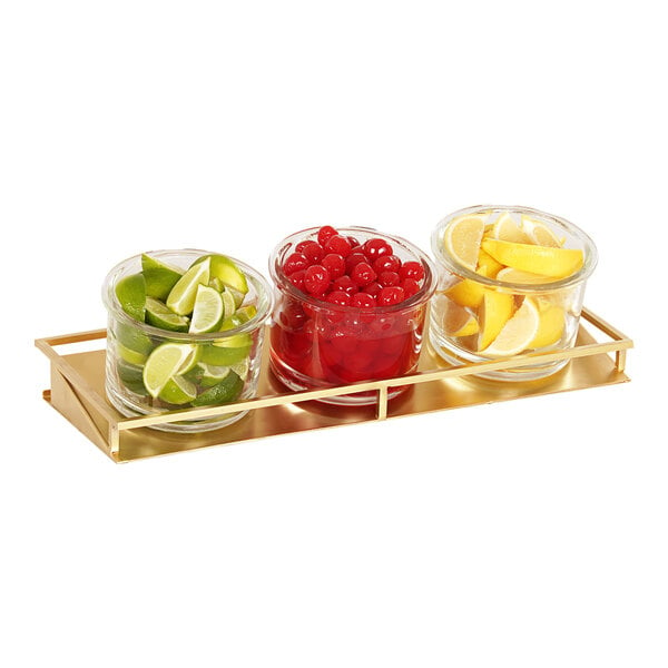 A Cal-Mil gold metal coffee condiment organizer with 3 glass jars filled with lemons, limes, and cherries.