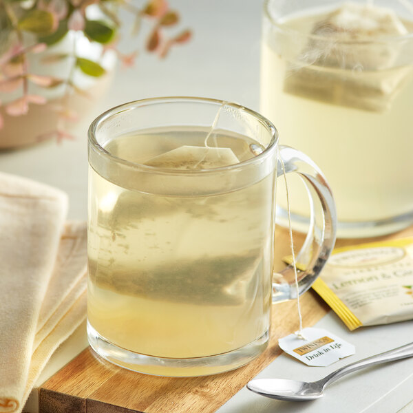 A glass mug of clear Twinings Lemon & Ginger tea with a tea bag on a wooden board.