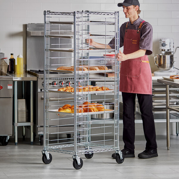 A person in a commercial kitchen is pushing a tall, metal wire rack on wheels that holds several trays of baked goods.