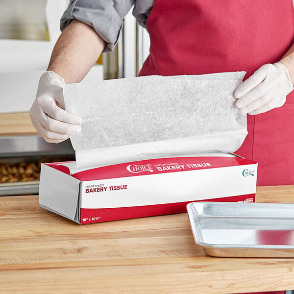 A person in a red apron holding a red and white box of Choice Customizable Interfolded Bakery Tissue Sheets.