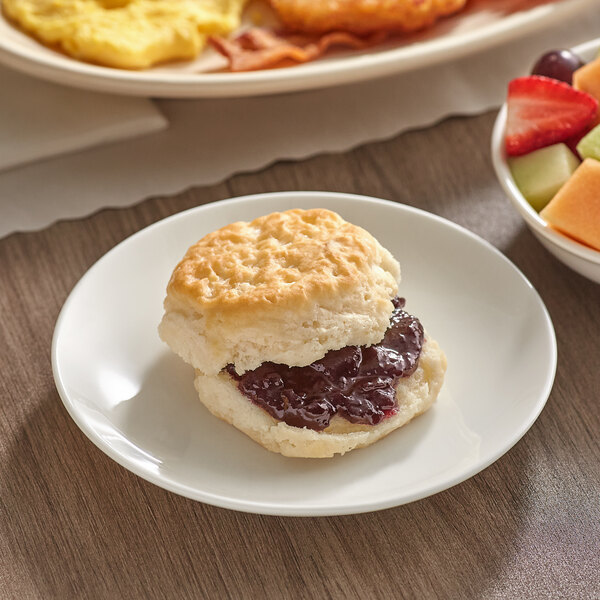 A Bakery Chef buttermilk biscuit with jam on a plate next to a bowl of fruit.