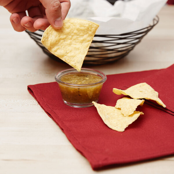 A hand holding a chip dipping into a clear plastic ramekin of salsa.