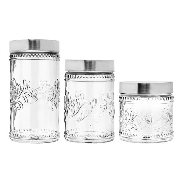 A group of three clear glass canisters with silver Fleur de Lis lids.