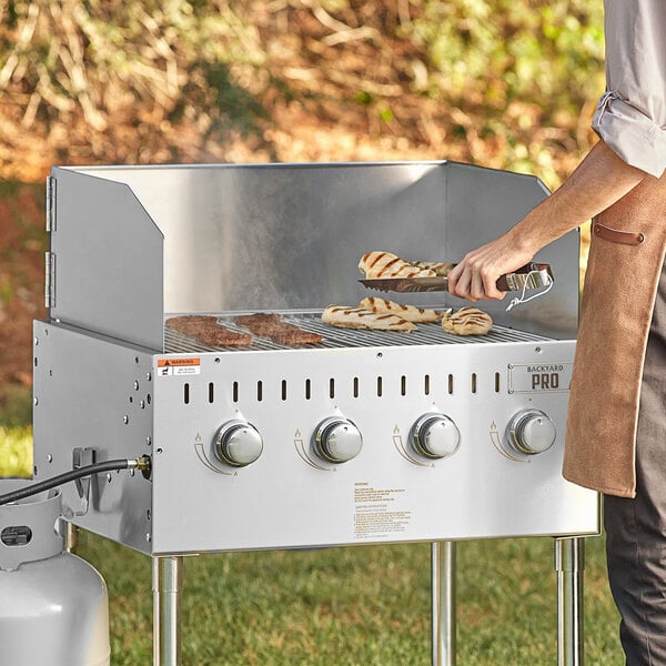 A person cooking food on a Backyard Pro stainless steel outdoor grill in the grass.