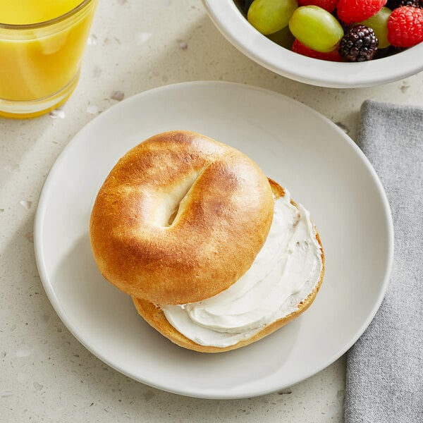 A Lender's plain bagel with cream cheese on a plate next to a bowl of fruit.