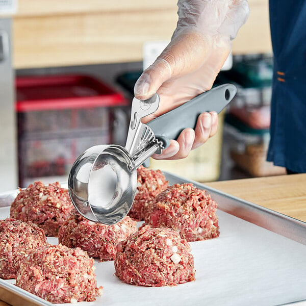 A person holding a Choice gray metal thumb scoop with a meatball in it.