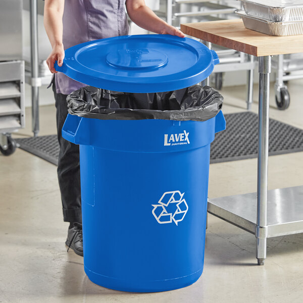 A woman standing in a school kitchen next to a blue Lavex recycling can with a blue lid.