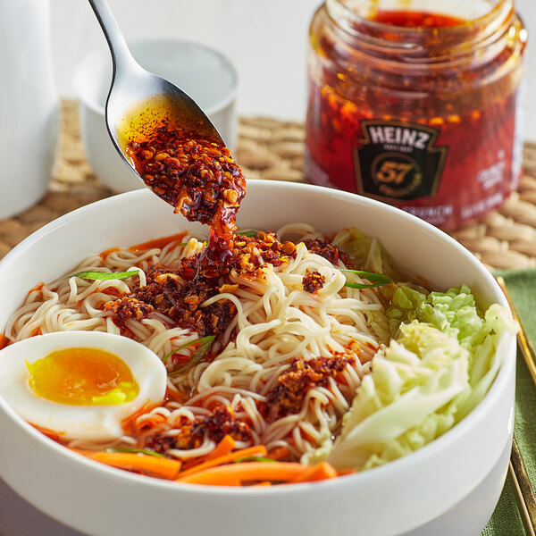 A bowl of noodles with Heinz Chili Pepper Culinary Crunch Sauce on a table.