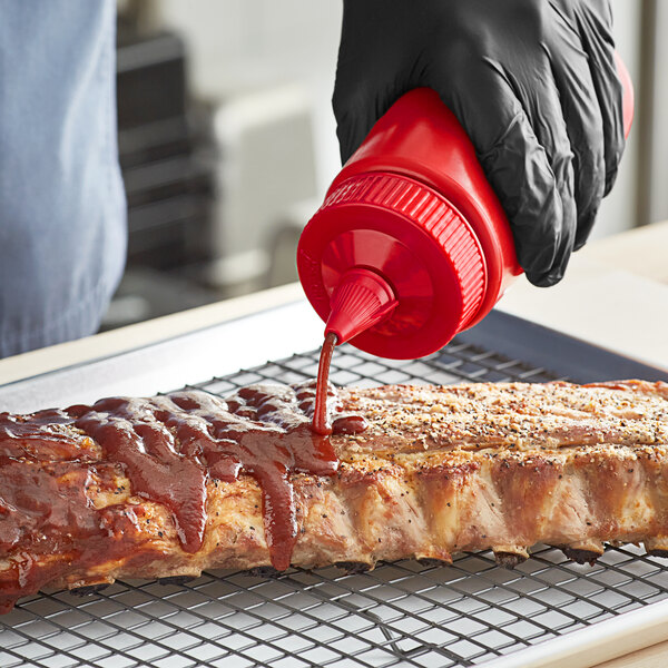 A hand using a Vollrath red bottle cap to pour barbecue sauce on a rack of ribs.