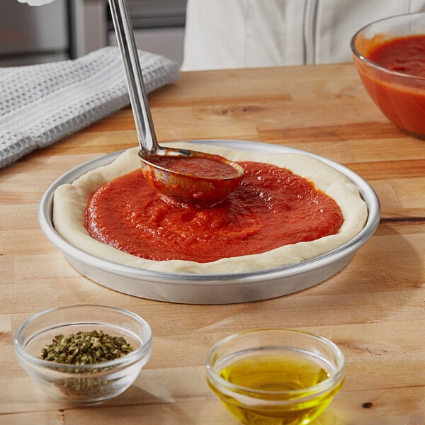 A person using a spoon to spread sauce on a pizza dough in a deep dish pizza pan.