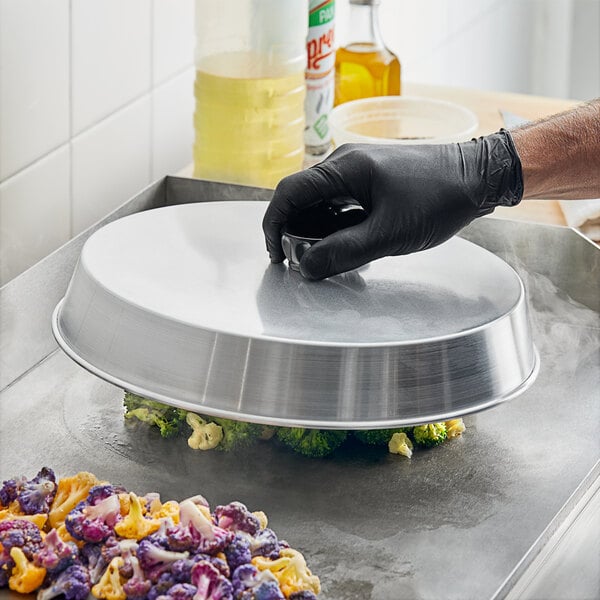 A person in black gloves using a Choice aluminum basting cover to cook broccoli on a pan.