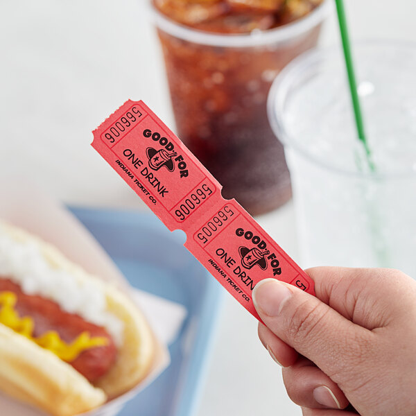 A hand holding a red "Good for One Drink" raffle ticket over a glass of soda.
