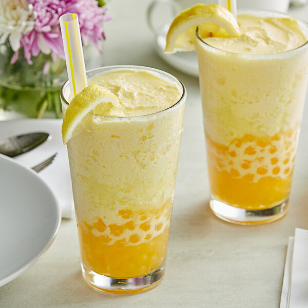 Two glasses of orange and yellow liquid with straws and lemon slices on a table.