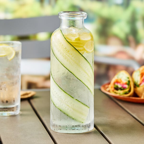 A Acopa clear glass water bottle with cucumber and lemon slices in a glass of water.