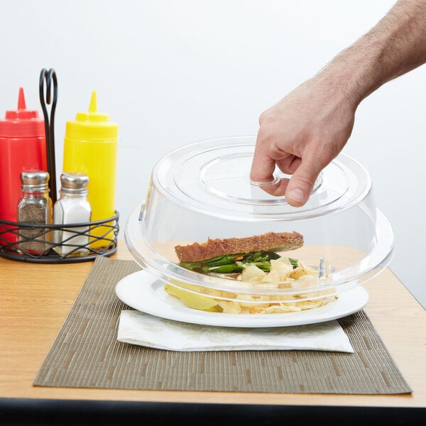 A hand reaching for a sandwich on a clear plate with a Cambro clear plate cover.