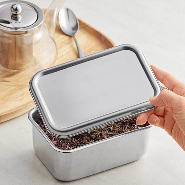 A hand holding a Matfer Bourgeat stainless steel container with a lid filled with tea and spices.