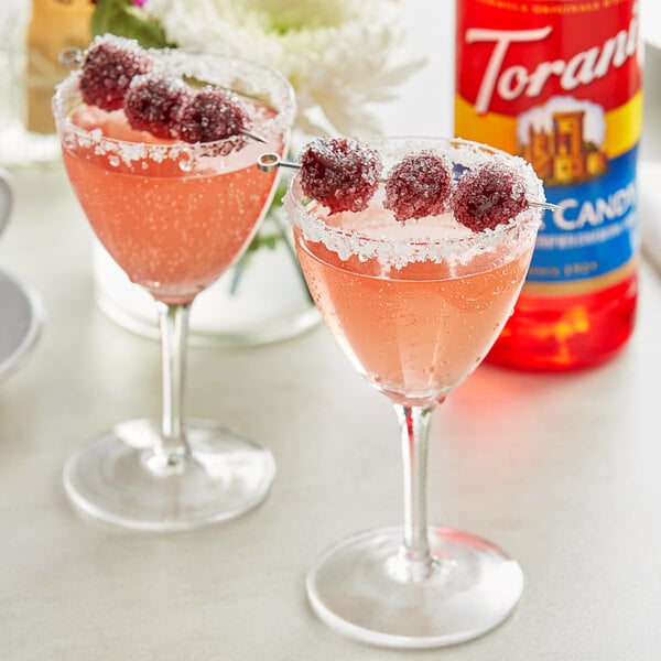 A glass of pink liquid with berries on top next to a bottle of Torani Sour Candy Flavoring Syrup.