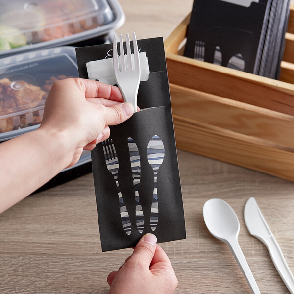 A person holding a black Dinex paper cutlery caddy with a fork and spoon inside.