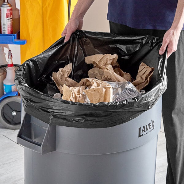 A man holding a black EcoStrong Plus trash bag over a trash can.