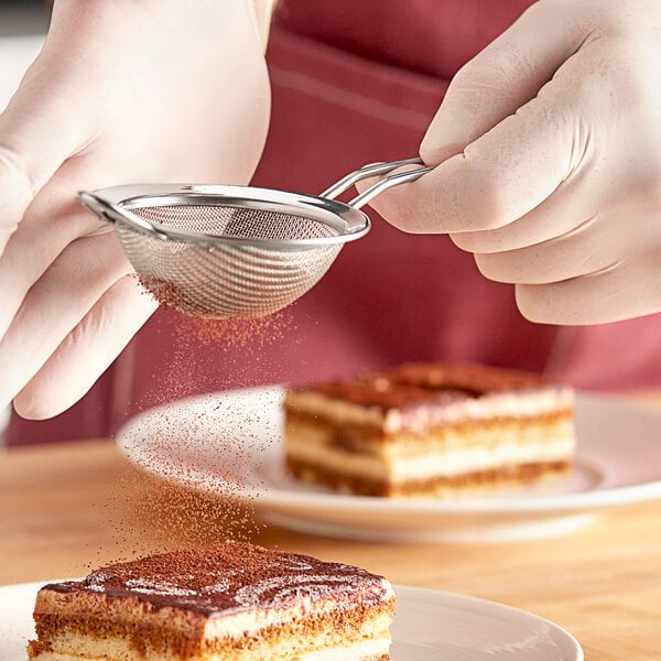 A person using a Matfer Bourgeat stainless steel fine mesh strainer to sprinkle powdered sugar over a piece of cake.