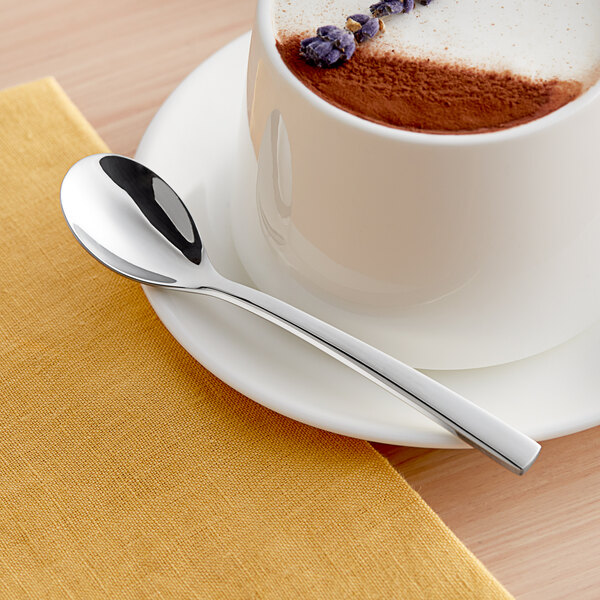 An Acopa stainless steel demitasse spoon on a saucer next to a cup of coffee.