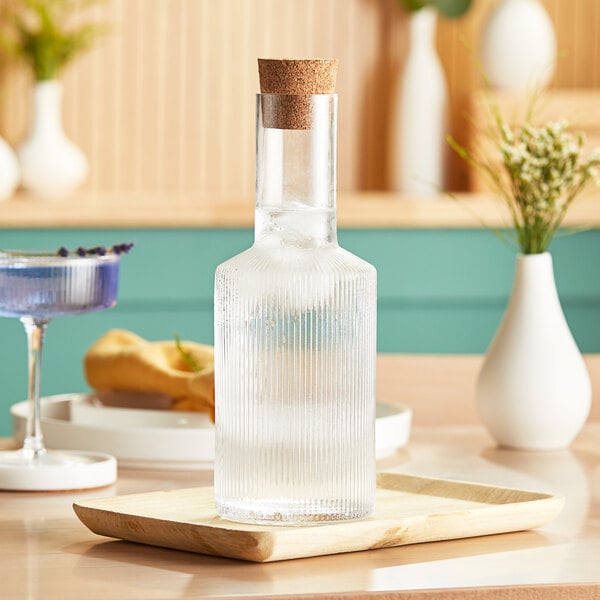 An Acopa clear glass bottle with a cork on a wooden tray.