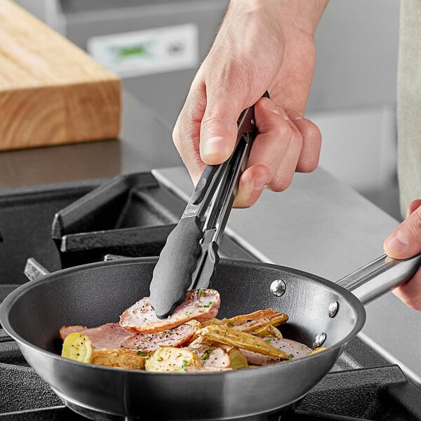 A person holding Choice 7" Silicone Tip Locking Tongs to a pan of food with meat and vegetables.