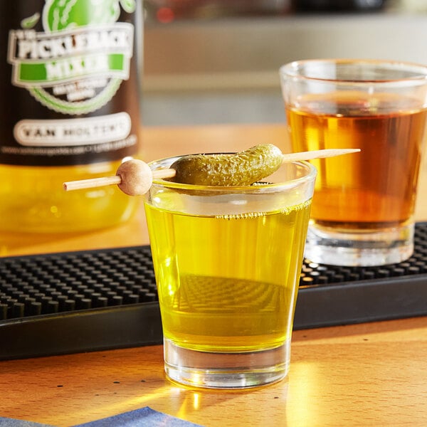 A glass of yellow liquid with a pickle on a toothpick.