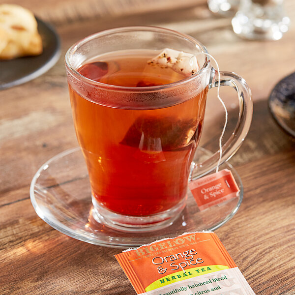 A glass cup of Bigelow orange & spice tea on a saucer with a Bigelow tea bag packet.