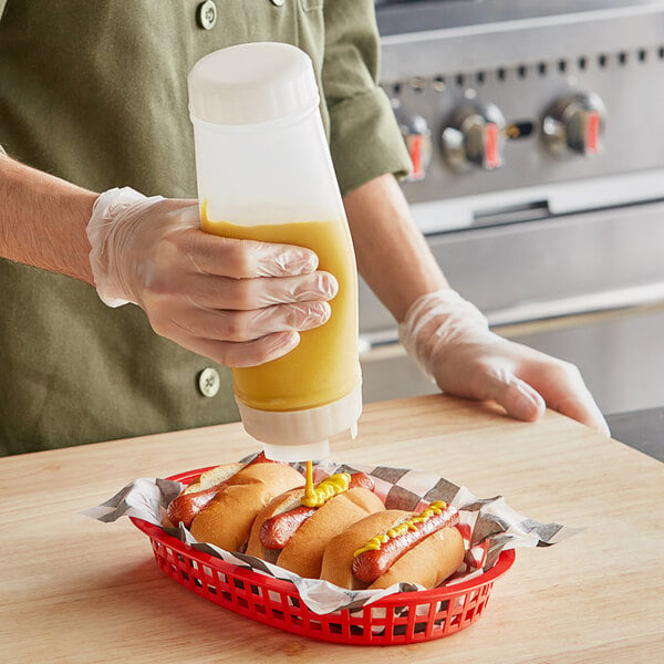 A person wearing gloves pouring yellow mustard from a San Jamar squeeze bottle onto a hot dog in a basket.