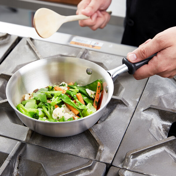 A person cooking vegetables in a Vollrath stainless steel saucier pan with a black handle.