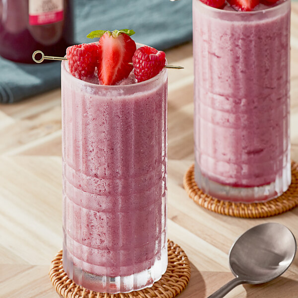 A close-up of a bottle of SHOTT Three Berry Real Fruit Flavoring Syrup with two glasses of pink smoothie with strawberries and raspberries.
