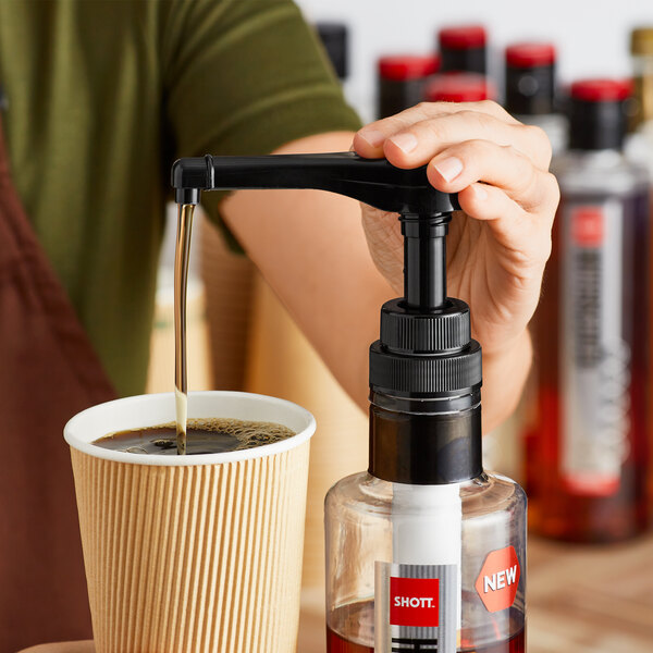 A person using a SHOTT syrup pump to pour liquid into a cup of coffee.
