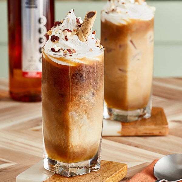 A glass of SHOTT hazelnut iced coffee with whipped cream and chocolate syrup with a spoon on a napkin.