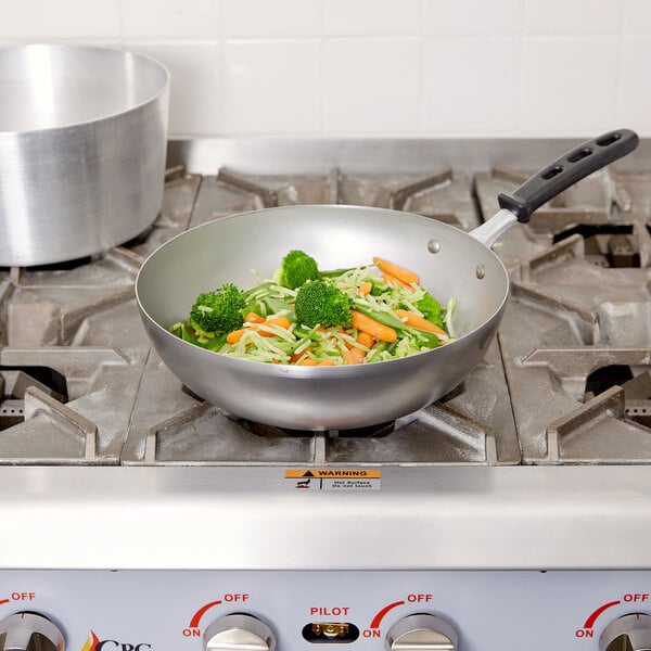A Vollrath carbon steel stir fry pan with broccoli and carrots on a stove.