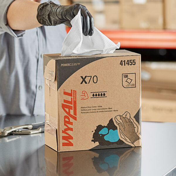 A person wearing black gloves cleaning a box of white WypAll industrial wipers.