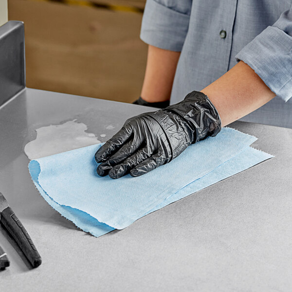 A person wearing black gloves cleaning a table with a blue WypAll X60 wiper.