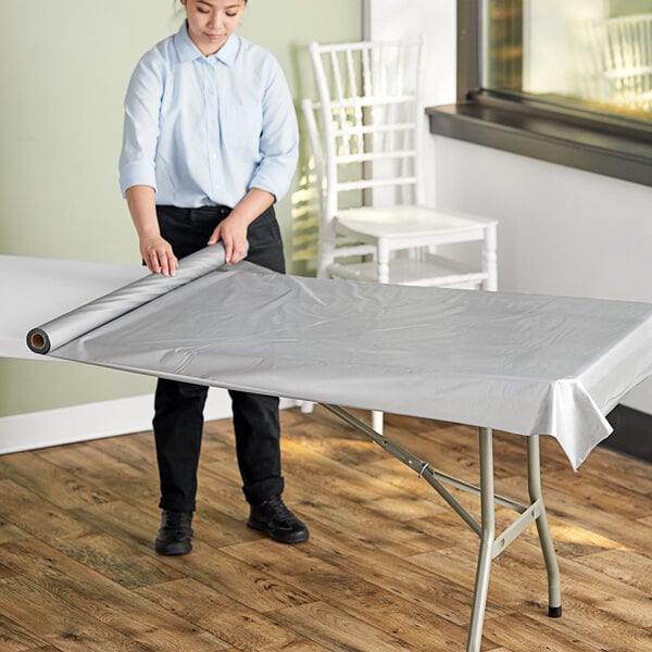 A woman rolling a metallic silver plastic table cover onto an outdoor table.