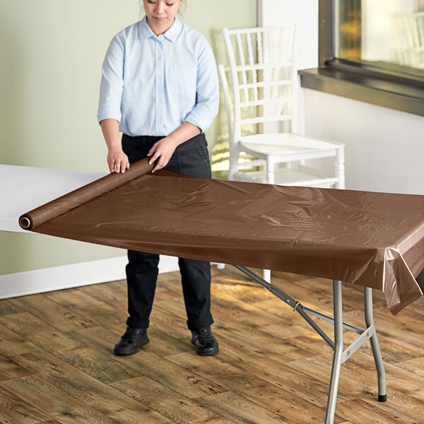 A woman rolling a chocolate brown plastic table cover on a table.