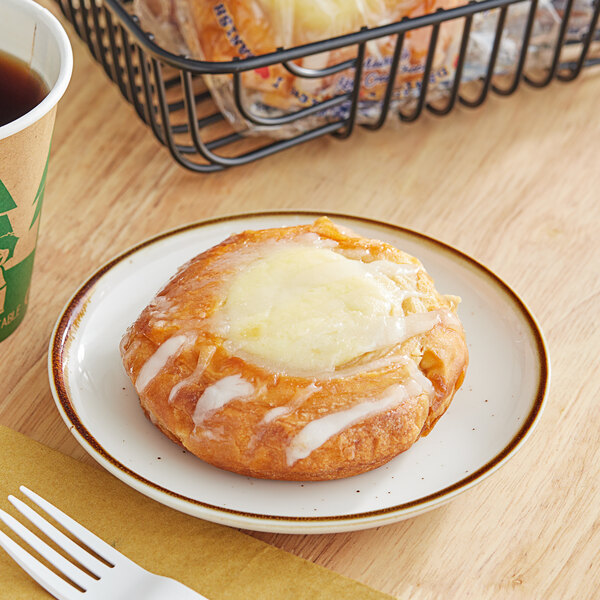 An Individually wrapped Ne-Mo's Bakery Cheese Danish on a plate next to a cup of coffee.
