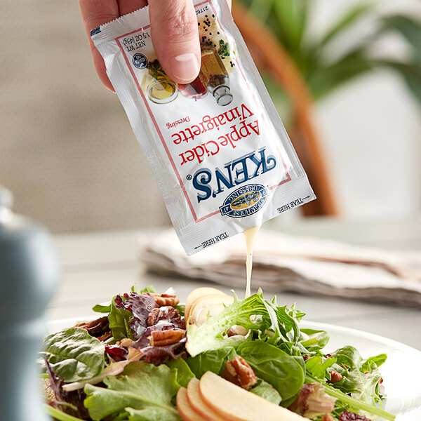 A hand pouring Ken's Foods Apple Cider Vinaigrette from a small packet onto a salad.