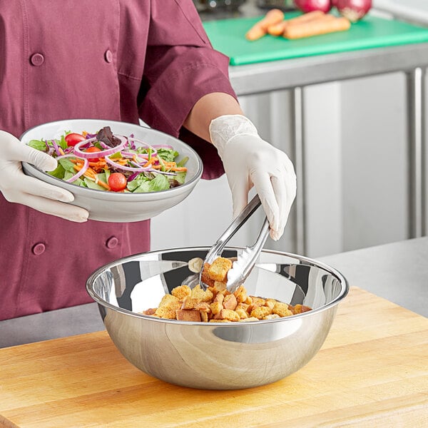 A person in a chef's uniform using a Choice stainless steel mixing bowl to prepare a salad with vegetables and onions.
