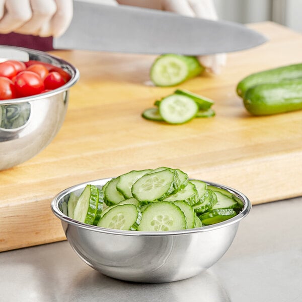 A person cutting cucumbers in a Choice stainless steel mixing bowl on a cutting board.