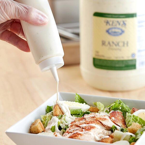 A person pouring Ken's Foods Ranch Dressing into a bowl of salad.