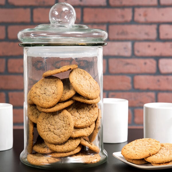 A Libbey glass jar filled with cookies on a table next to white coffee cups.