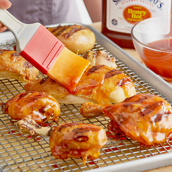 A person using a brush to apply Sweet Baby Ray's Hot Honey Wing Sauce to chicken on a grill.